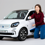 A friendly, professional female content creator, fully clothed in a modest, smart casual outfit. She is kneeling beside a vibrant children's electric car, pointing to a safety feature like a seatbelt or a parental remote control, explaining its function. Her expression is focused and informative. The setting is a brightly lit, clean studio with soft, neutral background elements, perhaps a workbench or shelving with other neatly arranged toy vehicles in the background, out of focus. professional photography, high detail, natural lighting, crisp focus, fully clothed, modest clothing, appropriate attire, professional dress, safe for work, appropriate content, family-friendly, perfect anatomy, correct proportions, natural pose, well-formed hands, proper finger count, natural body proportions.